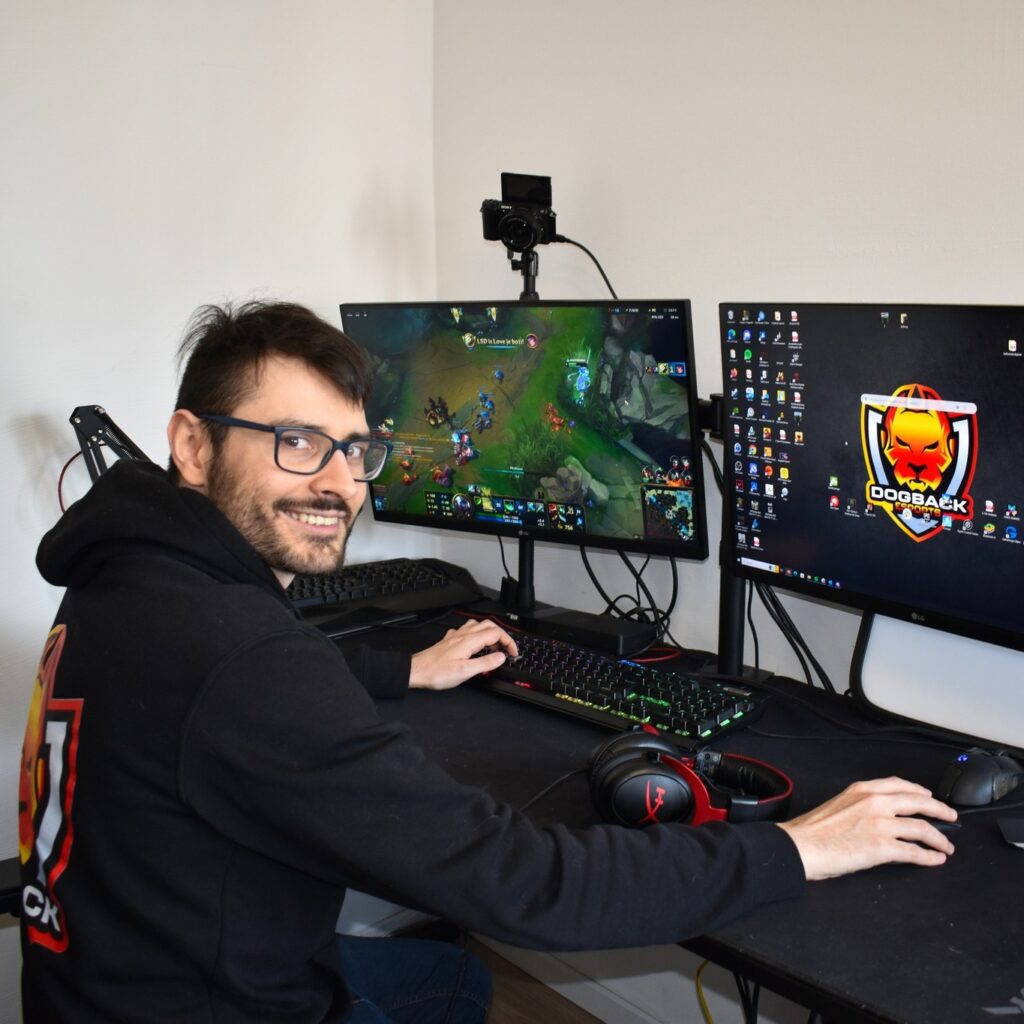 Lächelnder Mann spielt ein E-Sport-Spiel auf zwei Monitoren in einem Gaming-Setup, mit Dogback-Logo im Hintergrund.